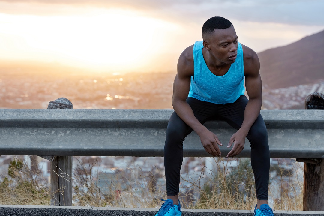 Athletic man rests after physical training, takes break on traffic sign, dressed in sportsclothes, thinks about sport competitions, magnificent dawn view